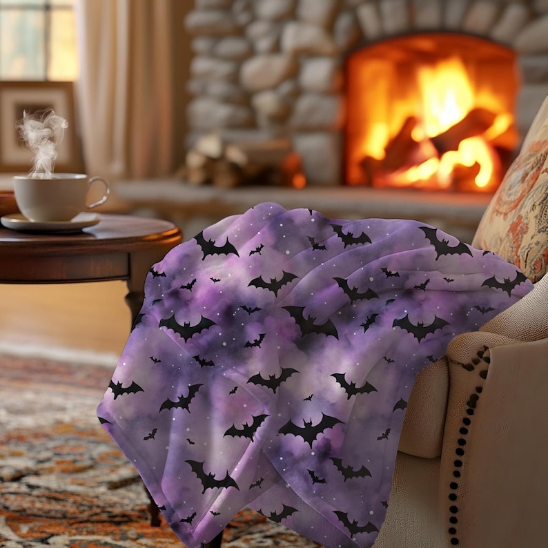 May include: A purple and white throw blanket with a pattern of black bats. The blanket is draped over a chair in front of a fireplace. The blanket has a watercolor effect.