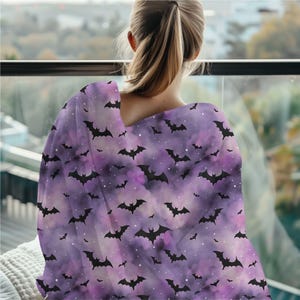 May include: A purple and lavender blanket with a pattern of black bats. The blanket is draped over a person's shoulders, with the bat design visible. The background is blurred, suggesting an indoor setting with a view.