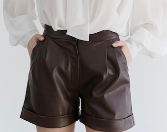 Op de afbeelding: Bruine leren shorts met een omgeslagen zoom. De shorts hebben een hoge taille en zakken. De shorts worden gecombineerd met een witte blouse met een strik.