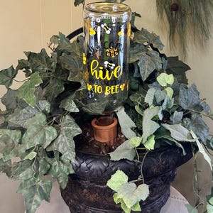 May include: A decorative bee-themed glass jar with the text "Welcome to our hive, the best place to bee" in yellow and black. The jar is set in a dark, ornate planter with green foliage.