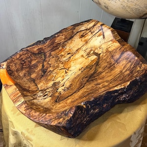 May include: A handcrafted wooden bowl with a natural, organic shape. The bowl features a rich, dark brown and tan wood grain pattern with a live edge. The interior has a smooth, polished finish, and the bowl is displayed on a golden tablecloth.