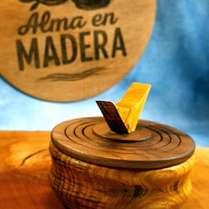 Puede incluir: Una caja de madera artesanal con tapa y un asa angular única. La caja está hecha de madera marrón clara y oscura, con patrones de vetas visibles. El fondo presenta un letrero de madera con el texto "Alma en Madera".