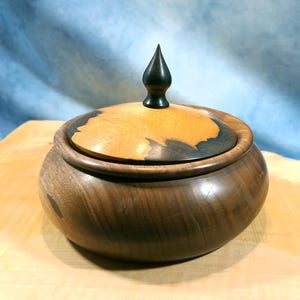Peut inclure: Un bol en bois artisanal avec un couvercle. Le bol est en bois brun foncé avec un couvercle en bois plus clair. Le couvercle a un bouton noir. Le bol est posé sur une surface en bois.