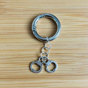 May include: A silver-colored keychain featuring a large circular clasp, several small connecting rings, and two miniature handcuffs. The handcuffs are detailed with small rivets and a locking mechanism.