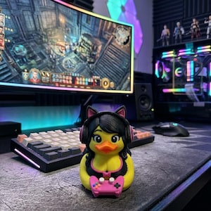 May include: A yellow rubber duck figurine featuring black hair, cat ears, and pink headphones. The duck wears a black top with pink trim and holds a small pink video game controller in its wings, positioned on a dark desk surface.