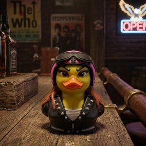 May include: A yellow rubber duck figurine styled as a biker, wearing a pink helmet with black goggles, long brown hair, and a black leather-style jacket with silver studs over a white shirt, featuring an orange beak and dark eye makeup.