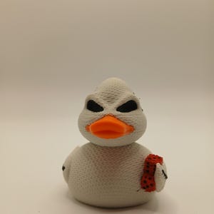 Puede incluir: Figura de pato blanco con ojos negros y pico naranja. El pato sostiene una bufanda roja con lunares negros. El pato tiene una superficie texturizada y está sobre un fondo blanco.