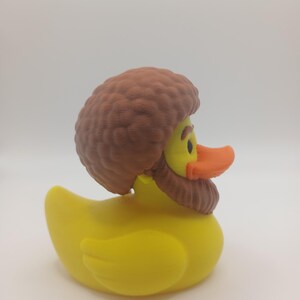 May include: A yellow rubber duck with a brown wig and beard. The duck has an orange beak and black eyes. The duck is a novelty item, likely for bath time or decoration.