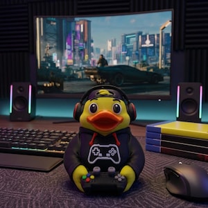 May include: A yellow rubber duck figurine wearing a black hoodie with a white game controller graphic and black headphones. The duck is holding a small black game controller in its wings, featuring a bright orange beak and large black eyes.