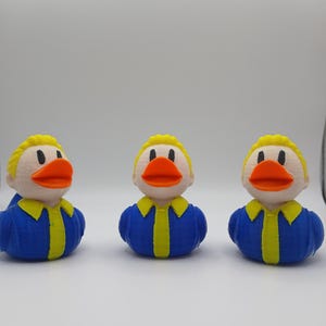 May include: Three 3D-printed duck figurines. Each duck has a yellow head, a bright orange beak, and a blue body with a yellow collar and tie. The ducks are arranged in a row against a white background.