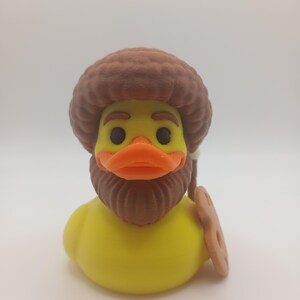 May include: A yellow rubber duck with a brown afro, beard, and orange beak. The duck is holding a brown cookie. This novelty item is a fun and unique gift.