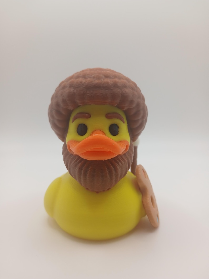 May include: A yellow rubber duck with a brown afro, beard, and orange beak. The duck is holding a brown cookie. This novelty item is a fun and unique gift.