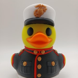 May include: A yellow rubber duck dressed in a U.S. Marine Corps uniform. The duck features a white hat with a gold emblem, a black and grey jacket with gold buttons, and an orange beak. The uniform has red detailing.