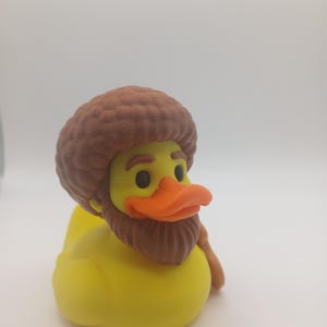 May include: A yellow rubber duck with a brown beard, hair, and a walking stick. The duck has an orange beak and black eyes. The duck is a novelty item.