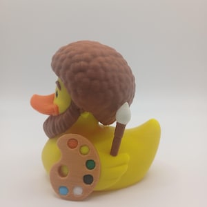May include: A yellow rubber duck with an orange beak, brown hair, and a matching beard. The duck is holding a wooden artist's palette with colorful paint and a paintbrush. The duck is a novelty item.