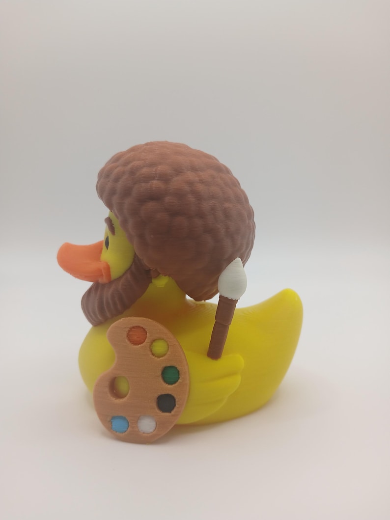 May include: A yellow rubber duck with an orange beak, brown hair, and a matching beard. The duck is holding a wooden artist's palette with colorful paint and a paintbrush. The duck is a novelty item.