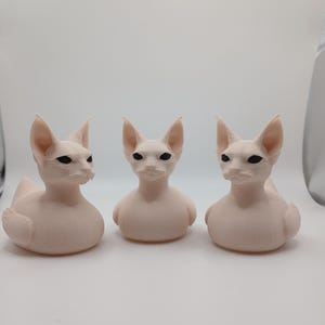 May include: Three light pink Sphynx cat figurines. Each figurine has large ears, black eyes, and a smooth, hairless appearance. The cats are in a sitting position, with their bodies rounded and their heads upright.