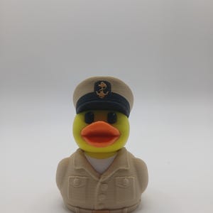 May include: A yellow rubber duck dressed in a tan naval uniform and hat with a black brim and gold anchor emblem. The duck has an orange beak and black eyes. A novelty item.