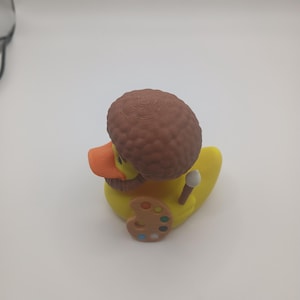 May include: A yellow rubber duck with an orange beak, brown hair, and a brown beard. The duck is holding a paintbrush and a painter's palette with various colors. The duck is a novelty item.