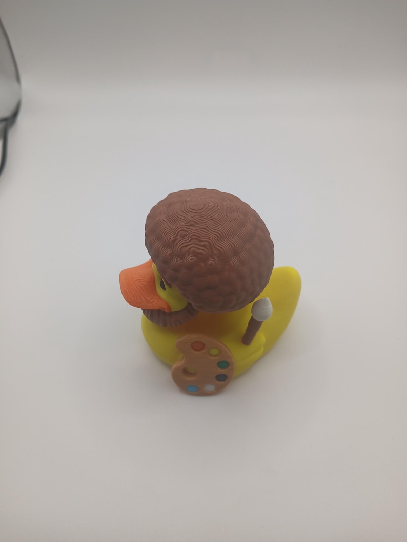 May include: A yellow rubber duck with an orange beak, brown hair, and a brown beard. The duck is holding a paintbrush and a painter's palette with various colors. The duck is a novelty item.