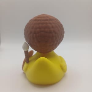 May include: A yellow rubber duck figurine with a brown, textured head and a small, white-tipped wand. The duck is viewed from the back, showcasing its rounded form and the details of its design.