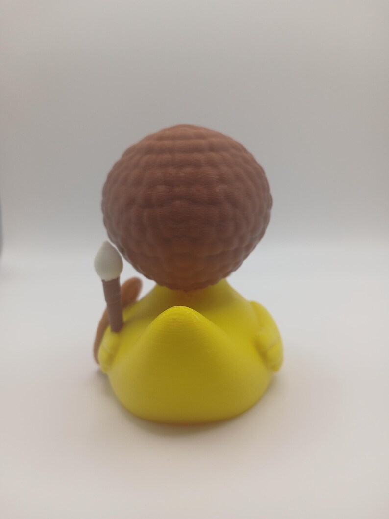 May include: A yellow rubber duck figurine with a brown, textured head and a small, white-tipped wand. The duck is viewed from the back, showcasing its rounded form and the details of its design.