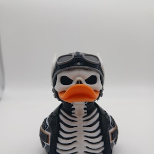 May include: A novelty rubber duck with a skeleton design. The duck is wearing a black leather jacket with a white rib cage design, a white helmet, and aviator goggles. The duck has an orange beak.