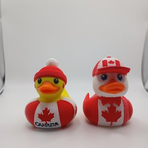Puede incluir: Dos patitos de goma con diseño de la bandera canadiense. Uno lleva un gorro a rayas rojas y blancas, el otro una gorra de béisbol roja. Ambos tienen picos amarillos y la hoja de arce canadiense.