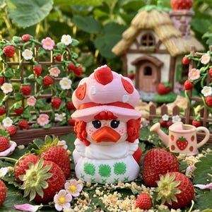 May include: A small duck figurine wearing a pink strawberry-patterned hat and a white dress with green cross-stitch details. The duck has orange features and red hair, positioned among fresh red strawberries and green leaves in a garden setting.
