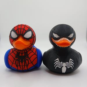 Puede incluir: Dos patitos de goma, uno diseñado como Spider-Man con detalles rojos, azules y blancos, y el otro como Venom, negro con detalles blancos. Ambos tienen picos naranjas y están sobre un fondo blanco.