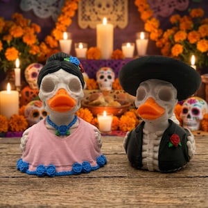 May include: Two duck figurines with skull-like faces and bright orange beaks. The left duck wears a pink dress with blue roses and a blue necklace. The right duck wears a black suit jacket, a red rose lapel pin, and a wide-brimmed black hat.