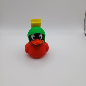 May include: A red rubber duck toy with a black face, orange beak, and white eyes. The duck is wearing a green helmet and a yellow square on top. The toy is a novelty item.