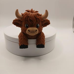Könnte beinhalten: Eine braune Highland-Kuh-Figur mit flauschigem Fell, großen Hörnern und schwarzen Hufen. Die Kuh hat große, runde Augen und sitzt auf einer weißen, runden Oberfläche. Die Figur ist ein bezaubernder Dekorationsartikel.