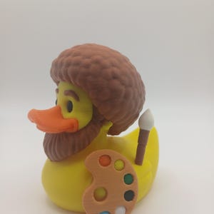 Puede incluir: Un patito de goma amarillo con peluca y barba marrones, pico naranja y una pequeña paleta de artista de madera con pintura colorida. Un pincel está metido detrás de la espalda del pato. El objeto es un artículo de novedad.