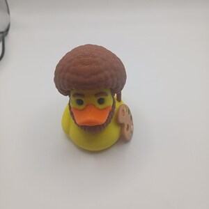 May include: A yellow rubber duck with a brown afro wig, a brown beard, and an orange beak. The duck has a small wooden artist's palette attached to its side. The duck is a novelty item.