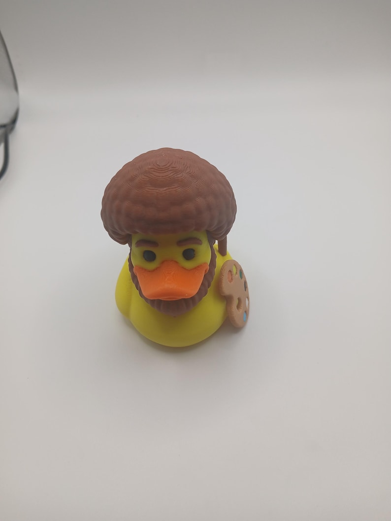May include: A yellow rubber duck with a brown afro wig, a brown beard, and an orange beak. The duck has a small wooden artist's palette attached to its side. The duck is a novelty item.