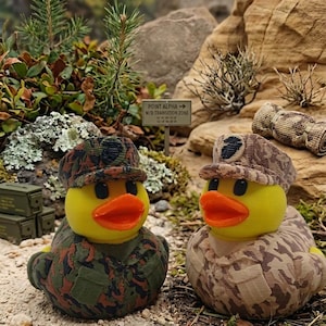 Peut inclure: Deux canards en caoutchouc jaunes vêtus d'uniformes de camouflage de style militaire avec des casquettes assorties. Un canard porte un motif forestier vert foncé et marron, l'autre un motif désertique beige. Ils ont des becs orange et des yeux noirs.