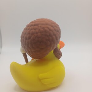 May include: A yellow rubber duck with a brown afro hairstyle and a small orange beak. The duck is wearing a white outfit and holding a brown stick. The toy is set against a white background.