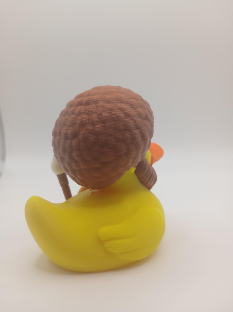 May include: A yellow rubber duck with a brown afro hairstyle and a small orange beak. The duck is wearing a white outfit and holding a brown stick. The toy is set against a white background.
