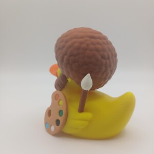 May include: A yellow rubber duck figurine with an orange beak and a brown afro wig. The duck is holding a paintbrush and a painter's palette with various colors. The figurine is a whimsical art-themed collectible.