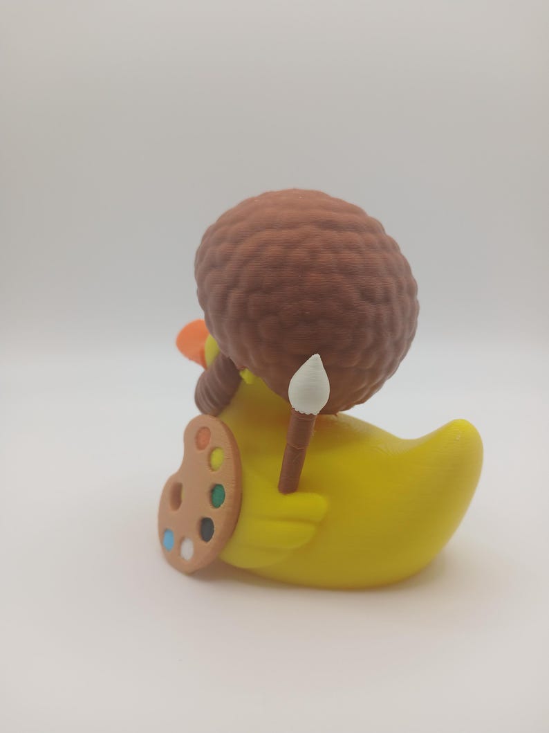 May include: A yellow rubber duck figurine with an orange beak and a brown afro wig. The duck is holding a paintbrush and a painter's palette with various colors. The figurine is a whimsical art-themed collectible.
