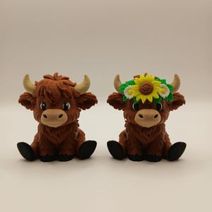 May include: Two brown Highland cow figurines. One cow has a flower crown with a yellow sunflower and green leaves. Both have tan horns, black hooves, and are sitting down. The figurines are on a white background.