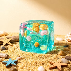 May include: A transparent turquoise resin cube containing various miniature seashells and small star-shaped glitter. The cube is clear with a glossy finish, showcasing a collection of pink, yellow, and white shells suspended within the material.