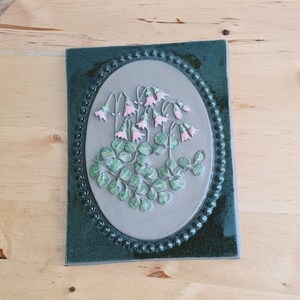 May include: A rectangular ceramic wall plaque featuring a dark green border with a beaded edge surrounding a light grey oval centre. The centre displays a raised relief design of green leaves and small, light pink bell-shaped flowers.