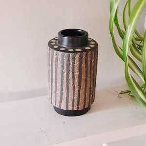 May include: A black and brown ceramic vase with a striped pattern and white dots around the top rim. The vase is sitting on a white shelf.