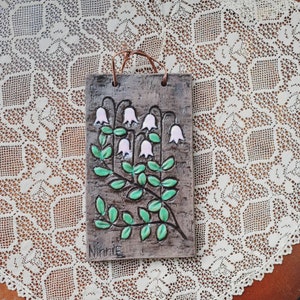 May include: A brown ceramic tile with a white and green floral design. The tile features a cluster of white bell-shaped flowers with green leaves. The tile is signed "Ninnie" in the bottom left corner.