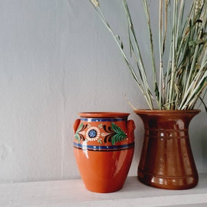Könnte beinhalten: Zwei Keramikvasen. Die erste ist ein leuchtendes Orange mit einem Blumendesign in Blau, Weiß und Grün. Die zweite ist eine dunkelbraune Vase mit einer weiten Öffnung. Getrocknete Weizenstängel befinden sich in der braunen Vase.