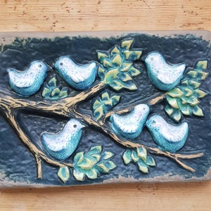 May include: A rectangular ceramic wall plaque featuring a relief design of five light blue birds perched on a textured brown tree branch with green leaves against a dark blue background. The plaque has a smooth, rounded border and a matte finish.