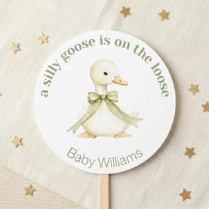 May include: A circular white sign featuring a watercolor illustration of a light yellow baby goose wearing a sage green ribbon bow around its neck. The text reads a silly goose is on the loose and Baby Williams in sage green lettering.