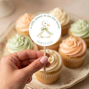 May include: A round white cupcake topper featuring a watercolor illustration of a white goose wearing a light green bow. The text reads a silly goose is on the loose and RHETT COY. The topper is attached to a thin wooden stick.